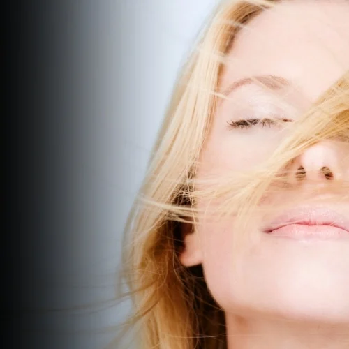 A woman with her eyes closed and hair moving in the wind against a soft grey background, representing the stillness, release, and return to self at the heart of Sonya Stattmann's work with women in the business arena.